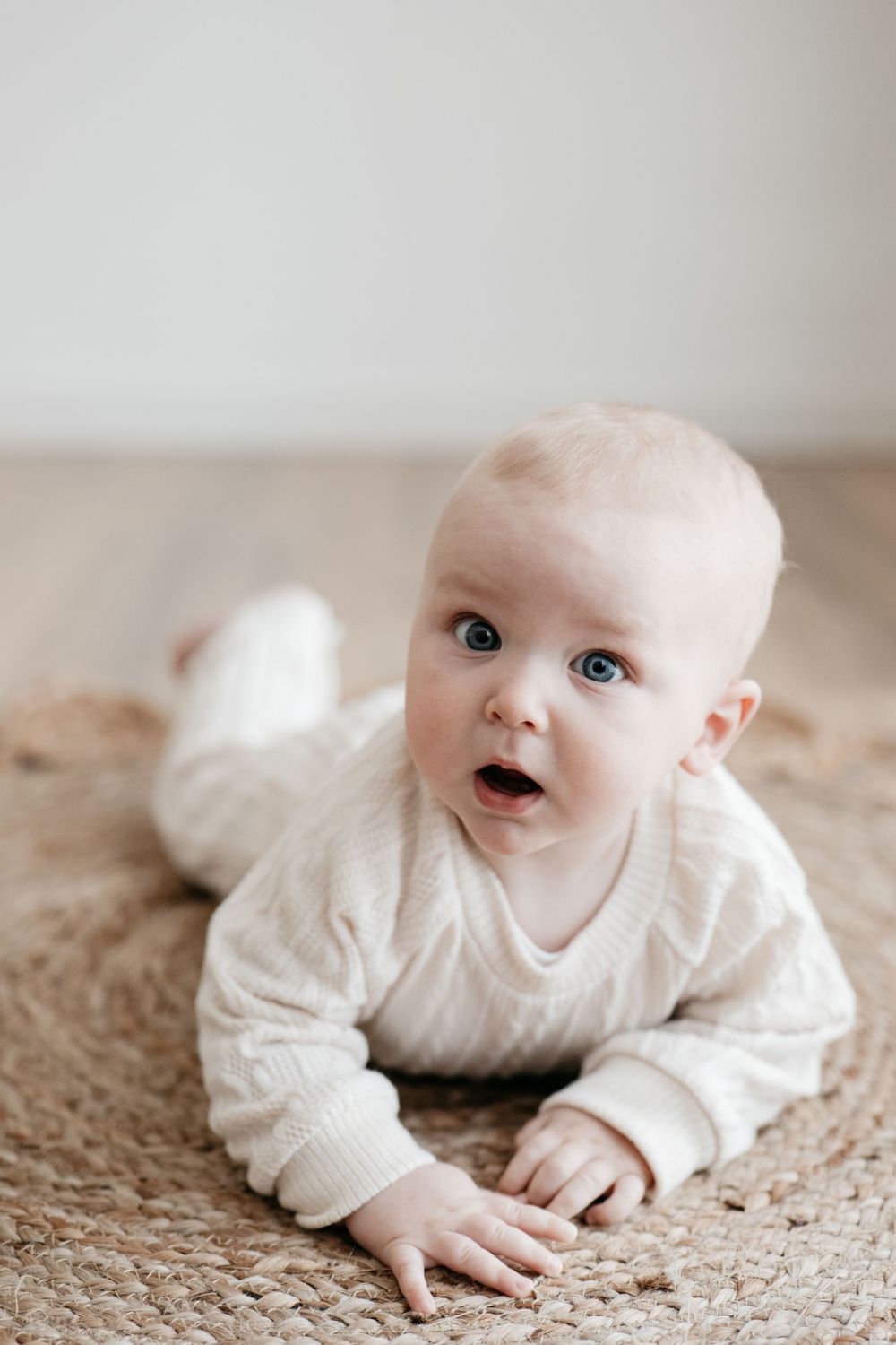 A baby in white knitwear lying on a beige carpet looking up with bright eyes.