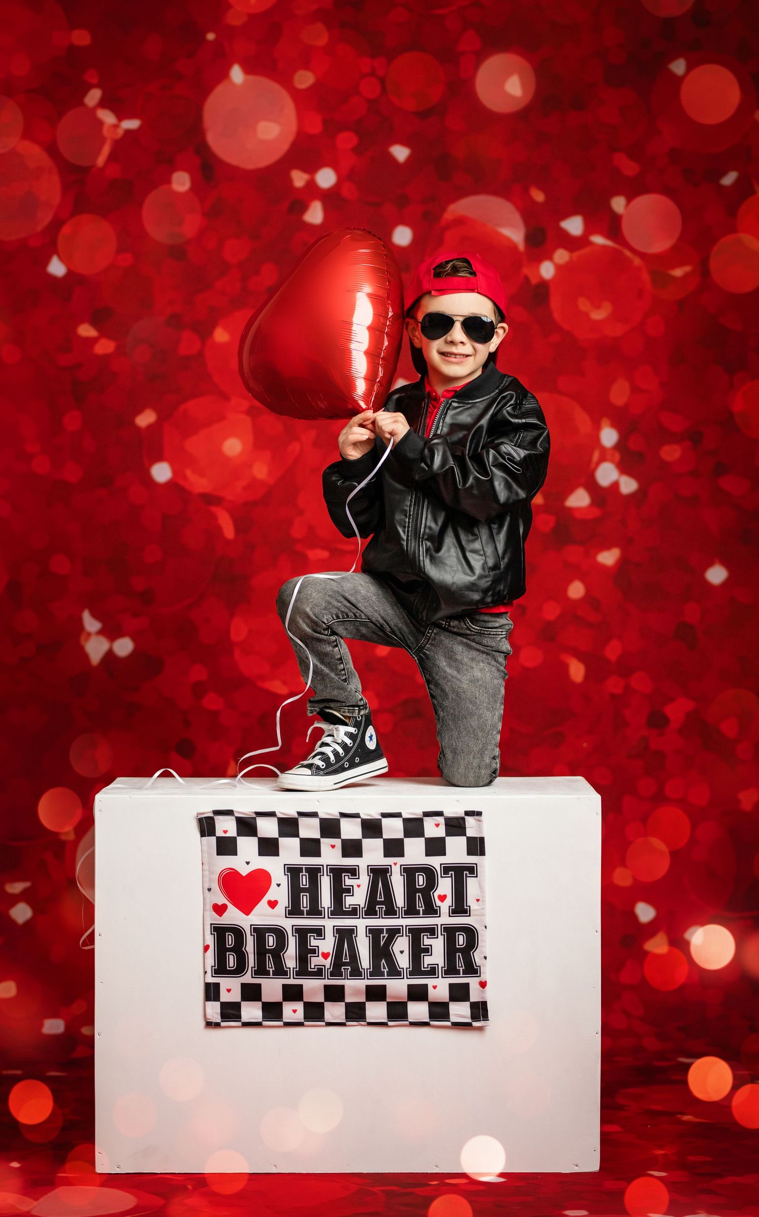 Young person in leather jacket and sunglasses poses with heart balloon against red bokeh backdrop for Valentine's theme.