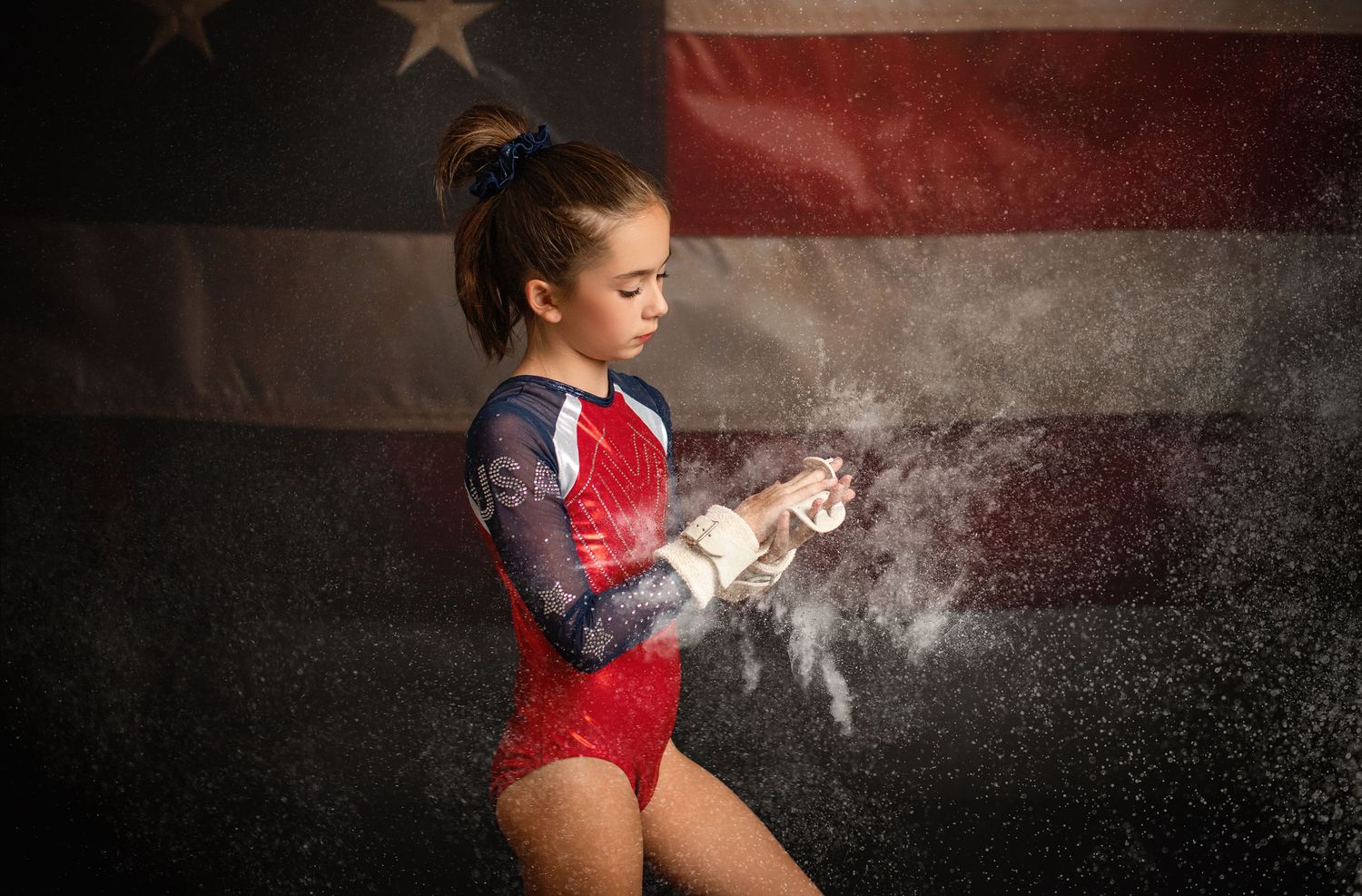 Gymnast in red leotard applies chalk powder to hands in front of an American flag background.