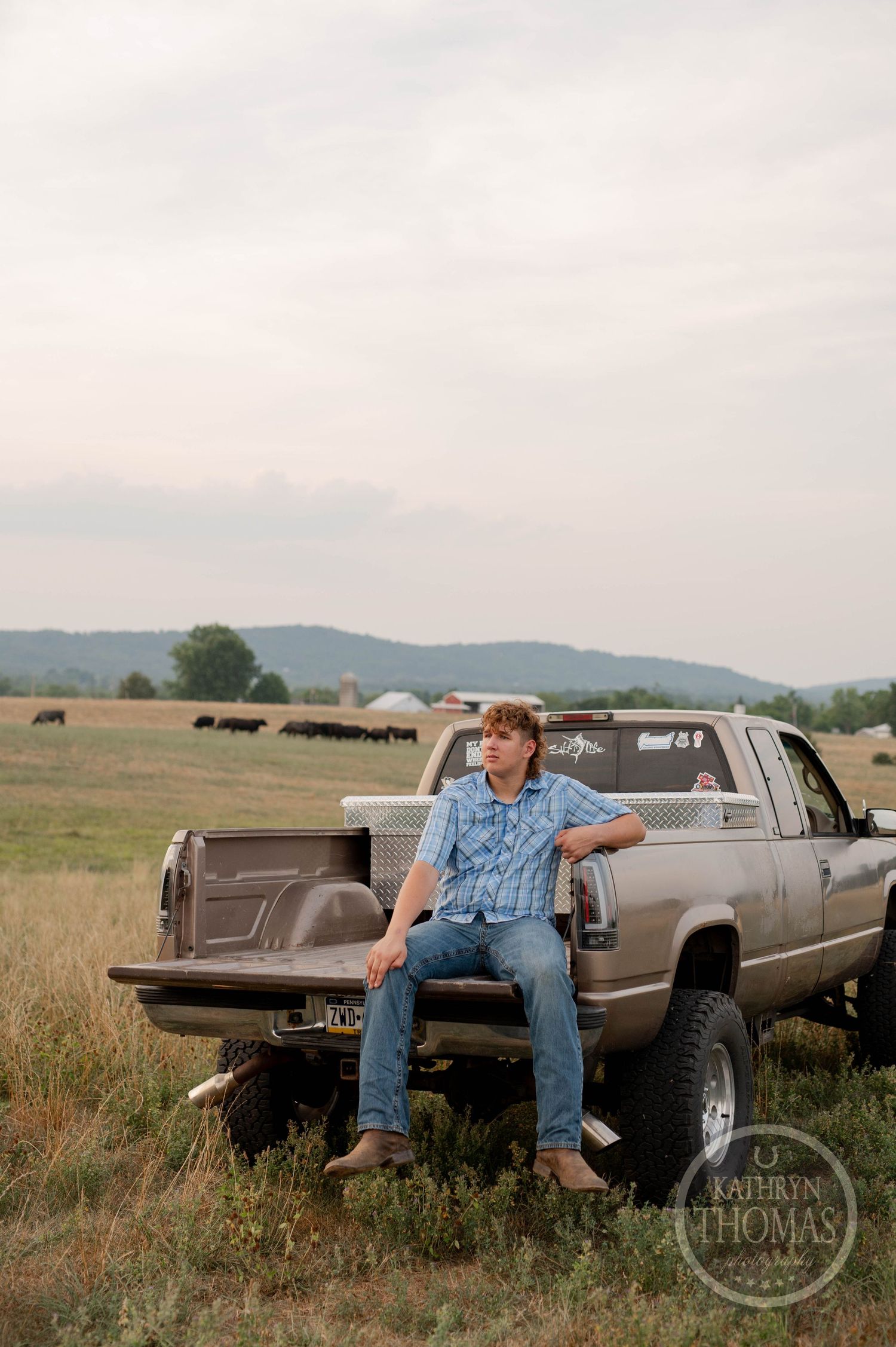 A person relaxes on the tailgate of a silver pickup truck with rolling hills in the background.