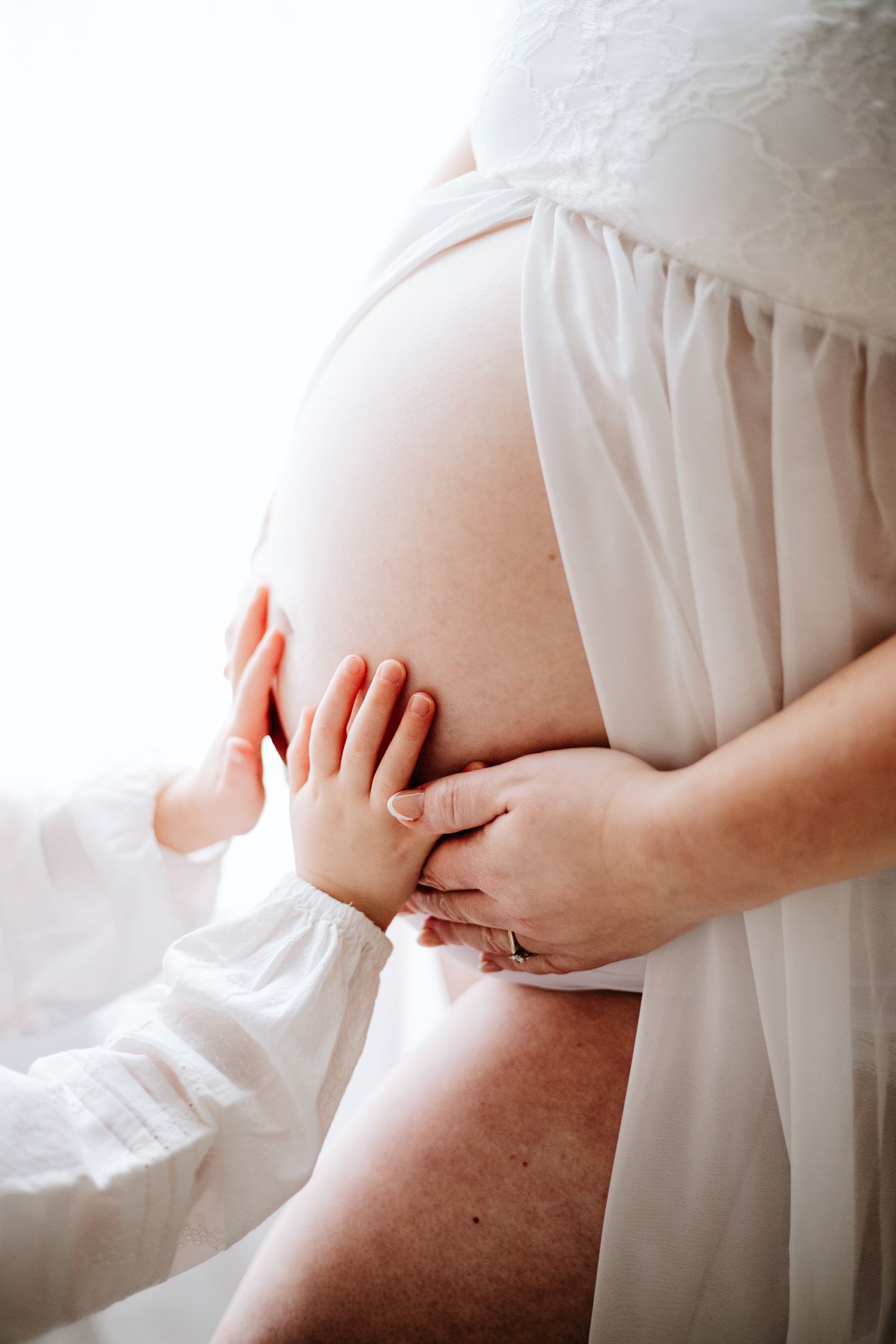 Small child's hands touch pregnant belly draped in white fabric in tender maternity portrait.