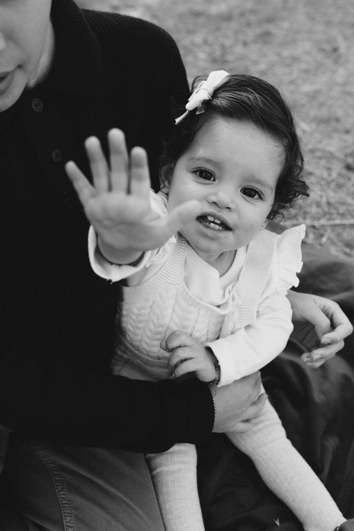 A small child in a white sweater waves at the camera while sitting on someone's lap in this black and white photo.