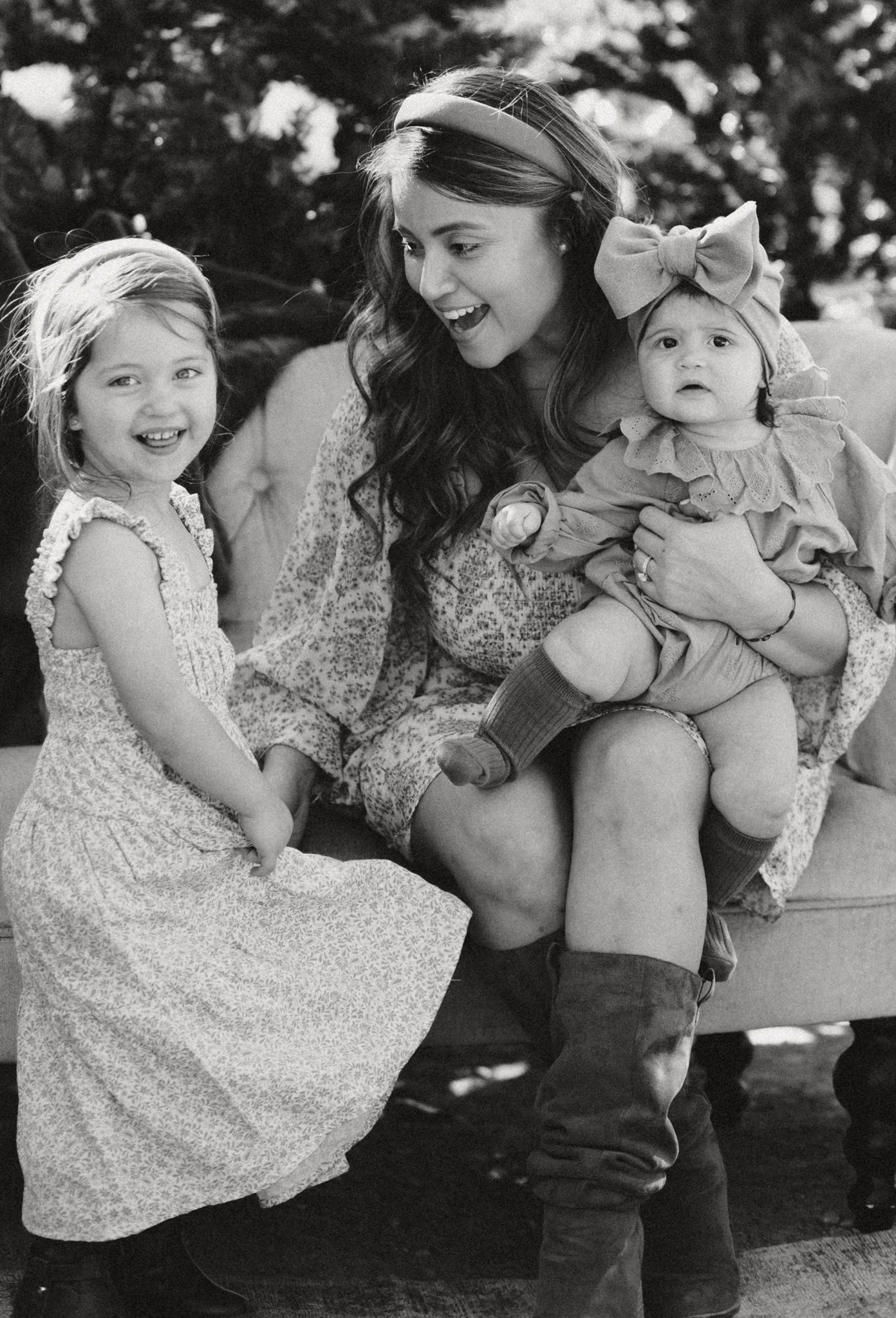 A black and white photograph shows three people sitting together outdoors in vintage-style dresses and clothing.