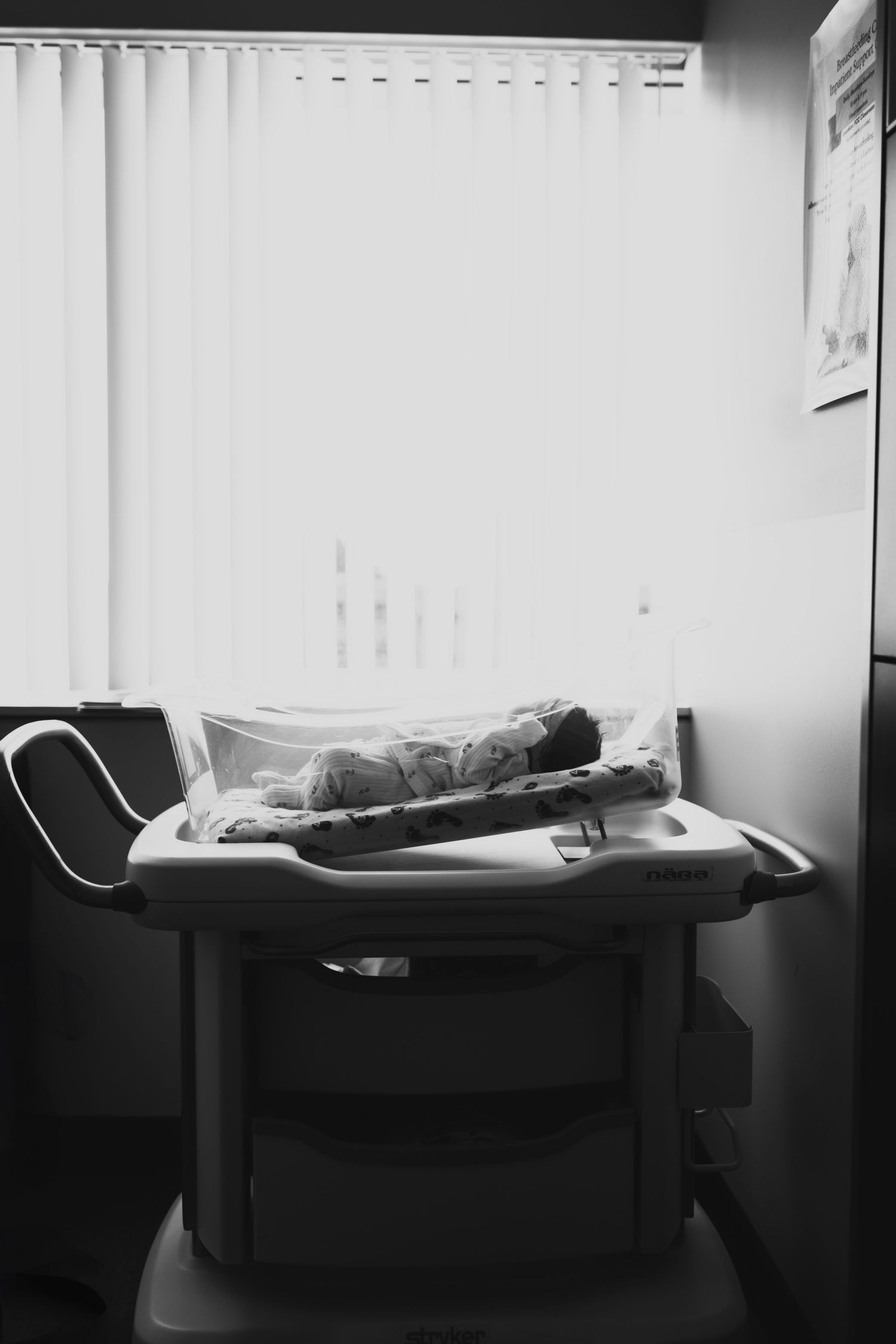 Black and white photo of a person lying down on a medical examination table in a clinical setting with vertical blinds.
