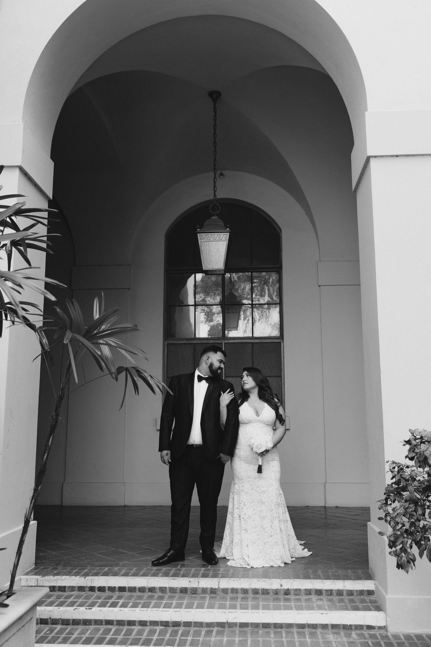 Black and white photo of a couple standing under an arched portico with hanging lantern and architectural details.