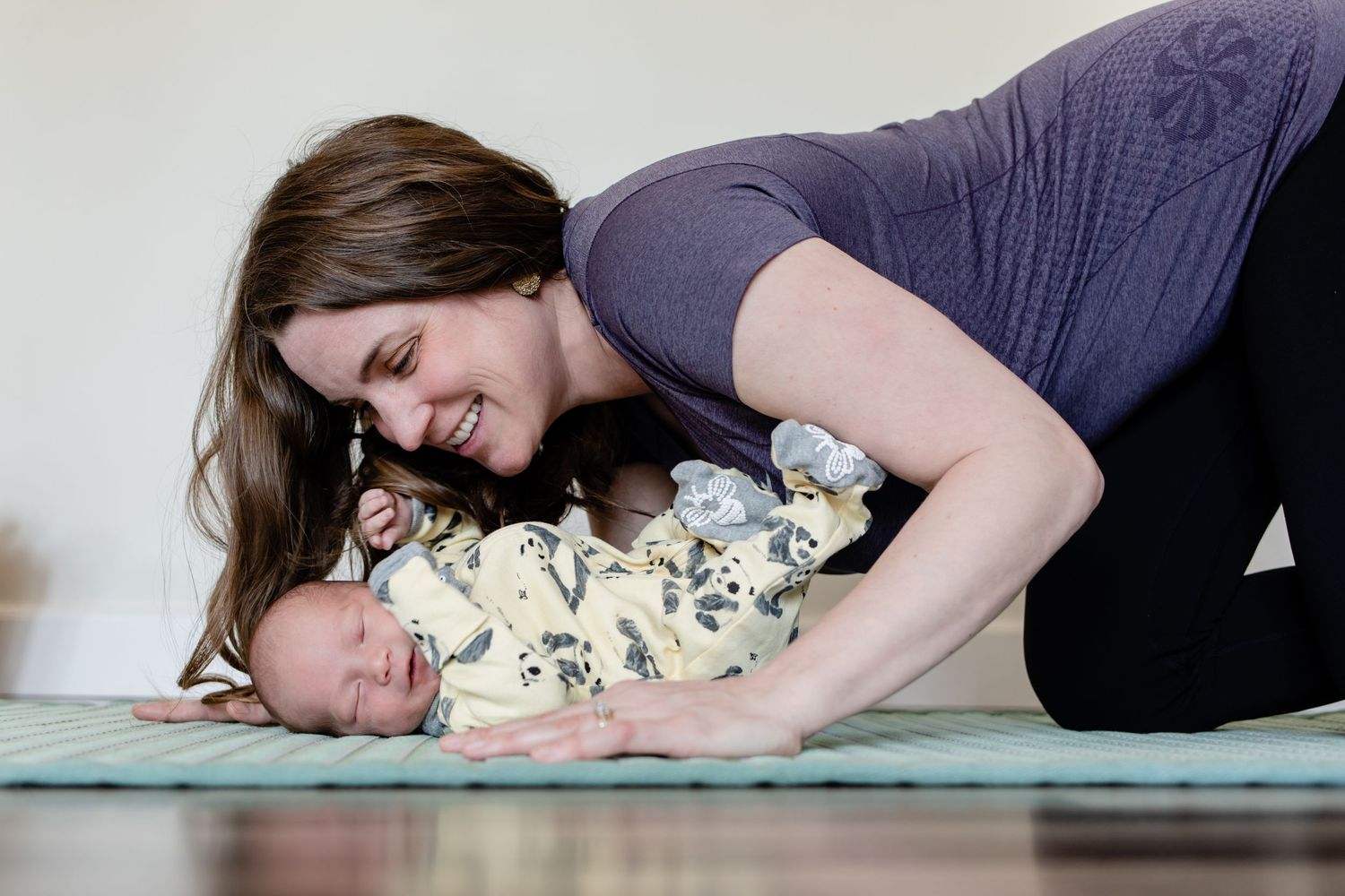 A mother lovingly embraces her sleeping baby who wears a white patterned onesie while lying on a blue mat together.