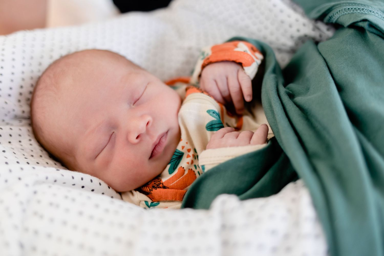 Sleeping newborn in green swaddle and fox print onesie rests on white knit blanket.