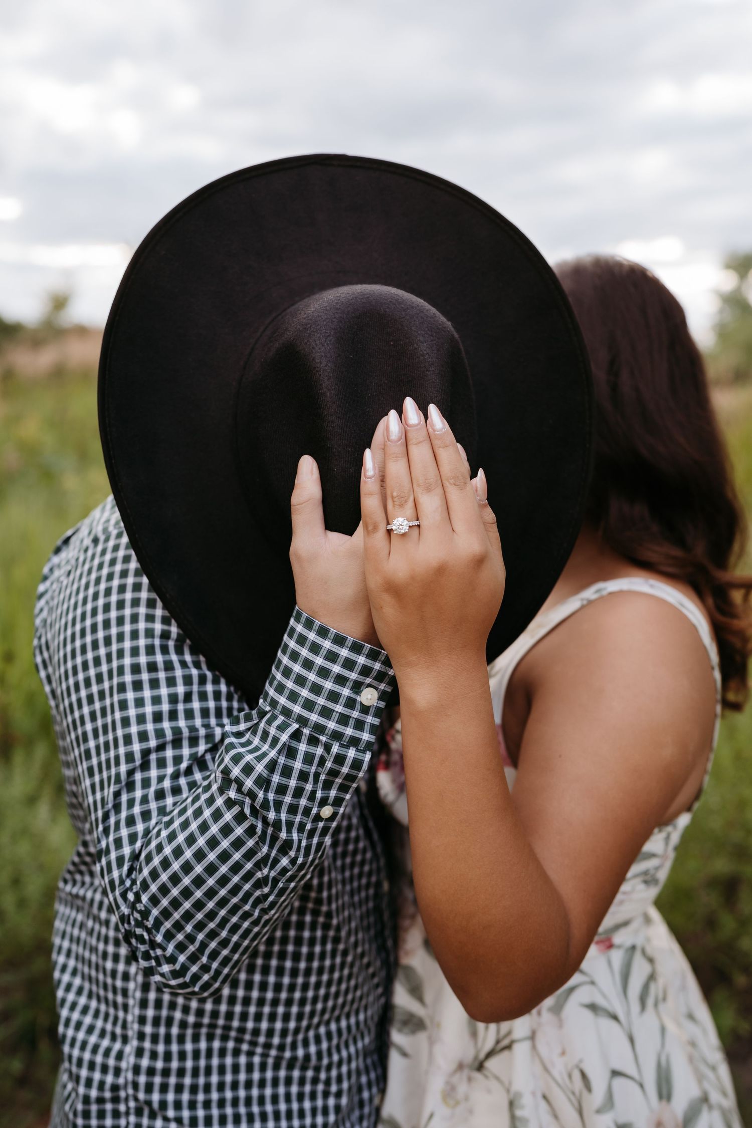 A couple shares an intimate moment in a field, with one wearing a black hat and checkered shirt.