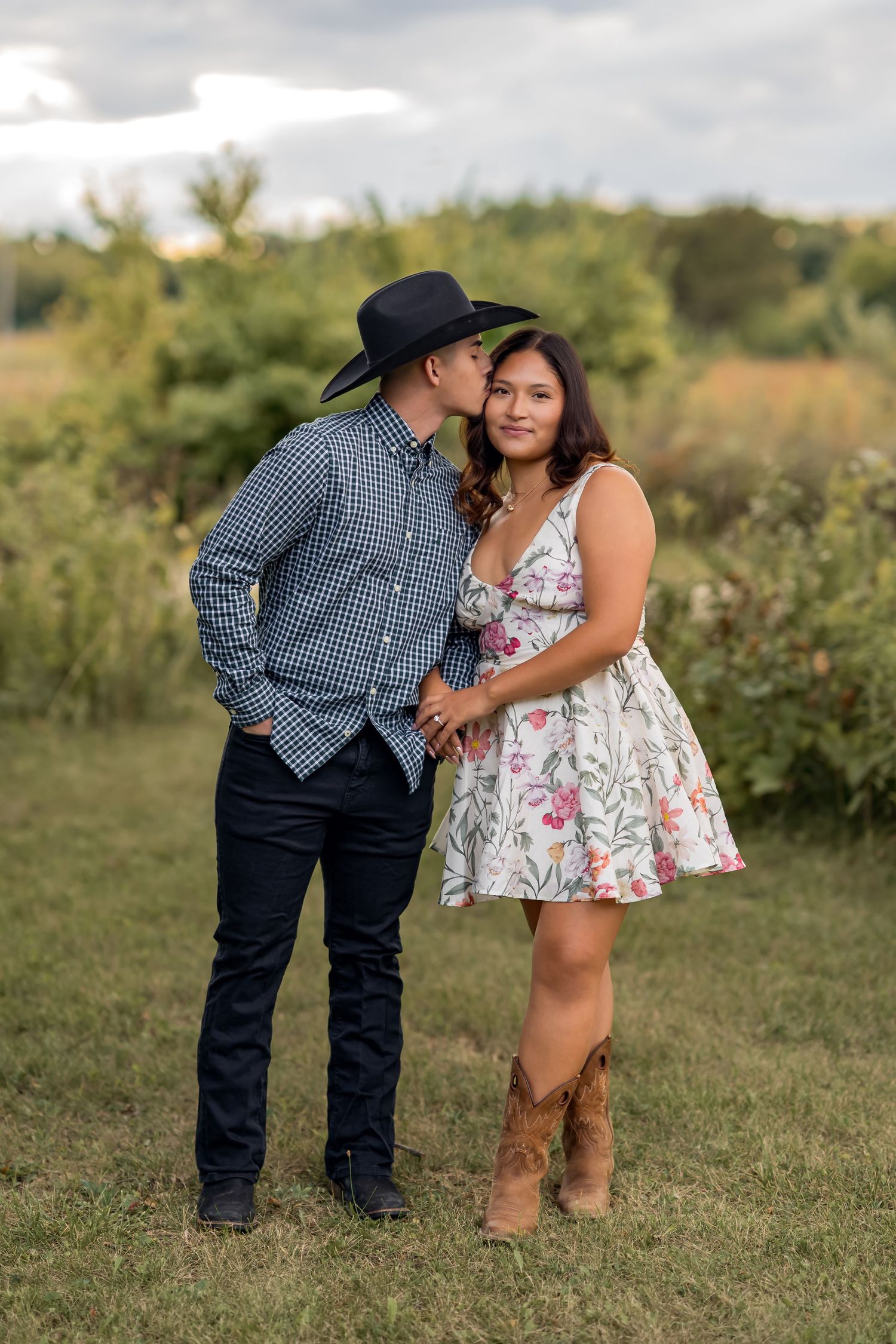 A couple shares a tender moment in a grassy field with one wearing a floral dress and another in western attire.