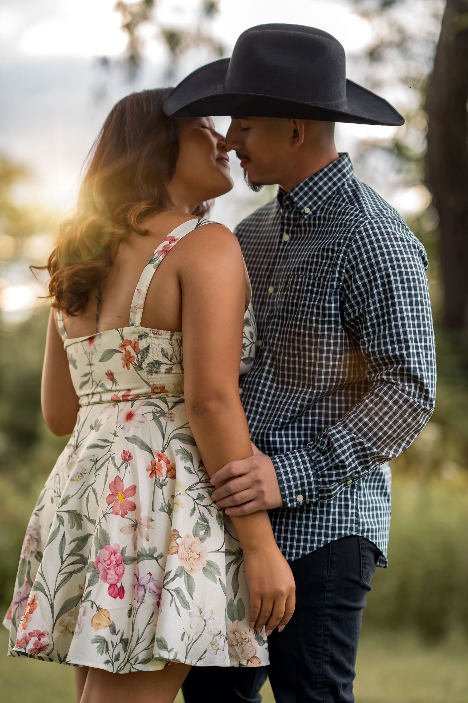 A couple in western attire, with a floral dress and plaid shirt, share tender moments in a sunlit outdoor setting.