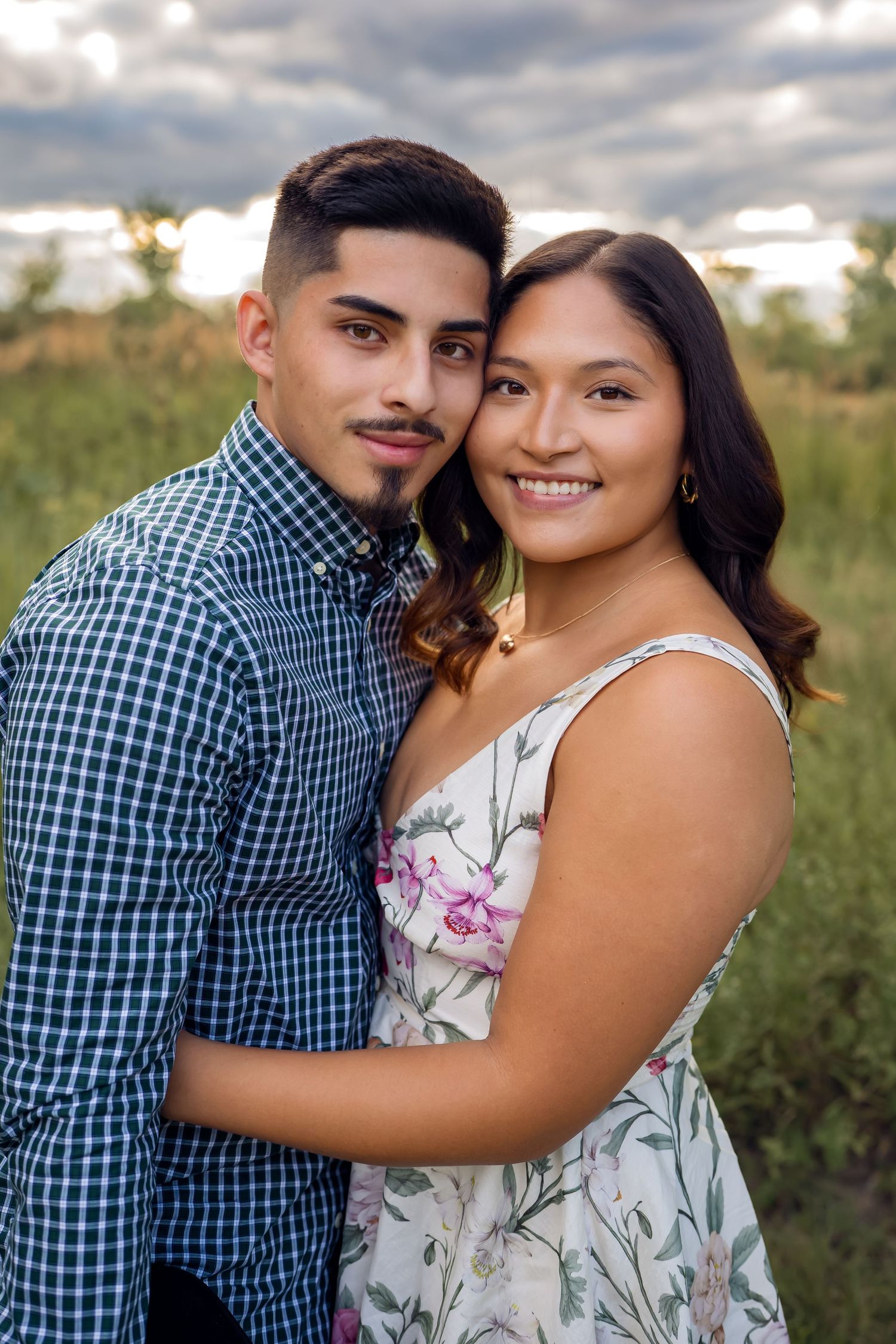 A couple embraces outdoors, with one in a checkered shirt and hat and the other in a floral dress.