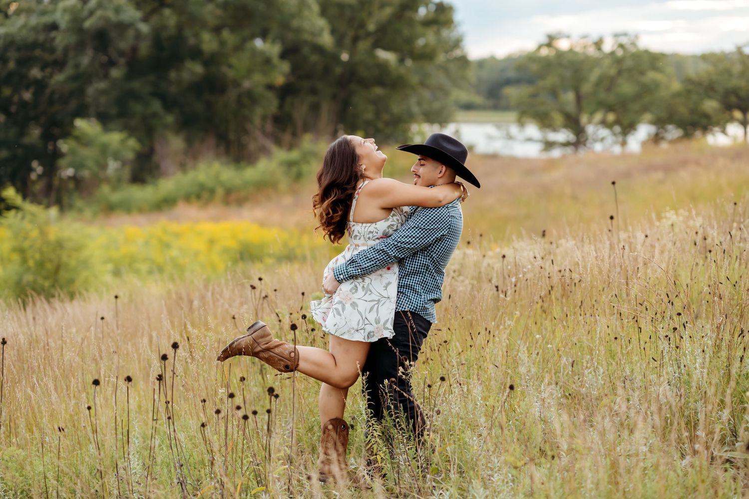 A couple embraces and dances playfully in a golden field during sunset, surrounded by tall grass and natural scenery.