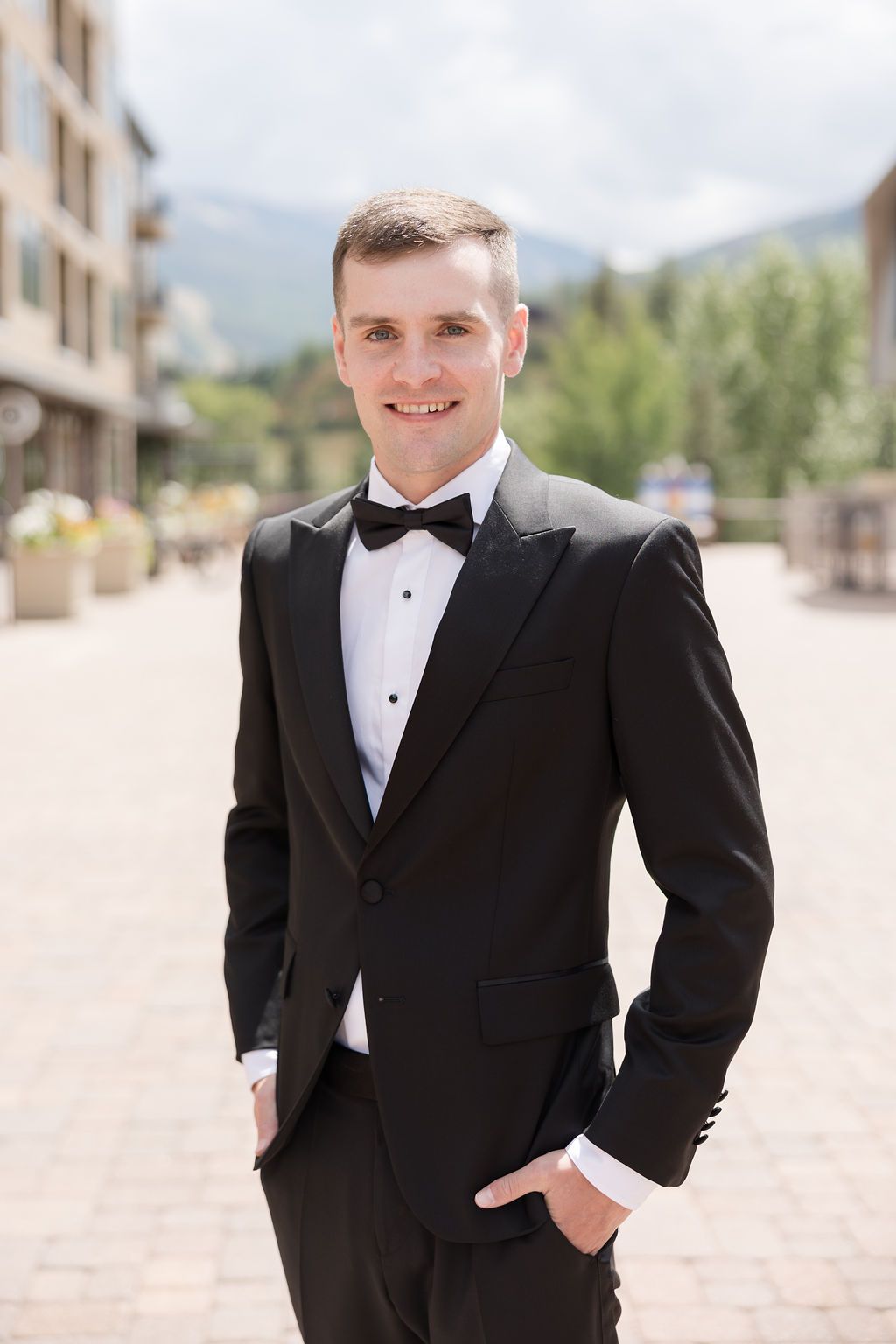 Groomsman in classic black tuxedo and bowtie poses outdoors at a wedding venue with buildings in background.