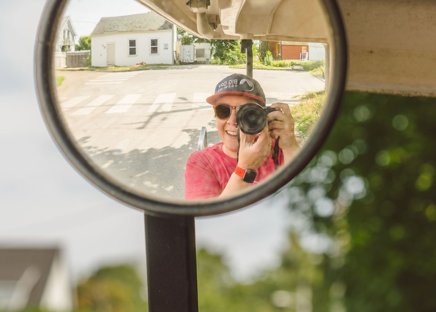 A reflection in a side mirror shows a photographer wearing a red shirt and sunglasses taking a picture.
