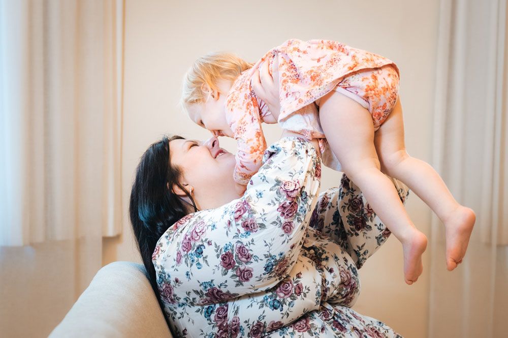Kukkamekkoiset äiti ja lapsi valokuvausstudion sohvalla, jossa äiti nostaa lastaan käsien varassa ilmaan.