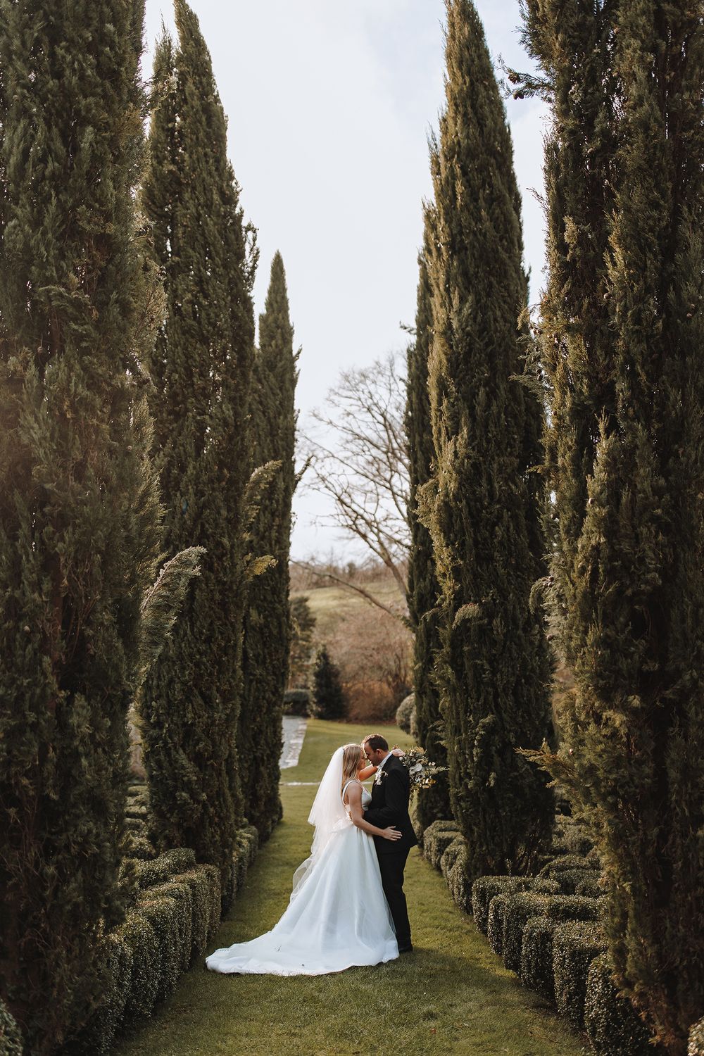 A romantic wedding couple poses between tall cypress trees in a majestic garden pathway.