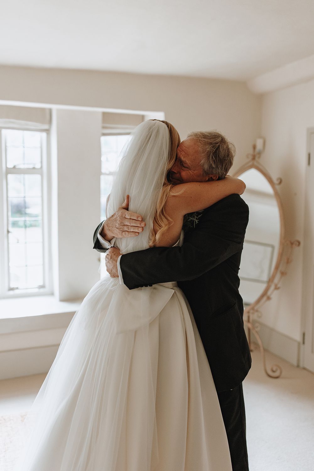 A tender embrace between a bride in a white wedding gown and someone in a black suit in a bright, sunlit room.