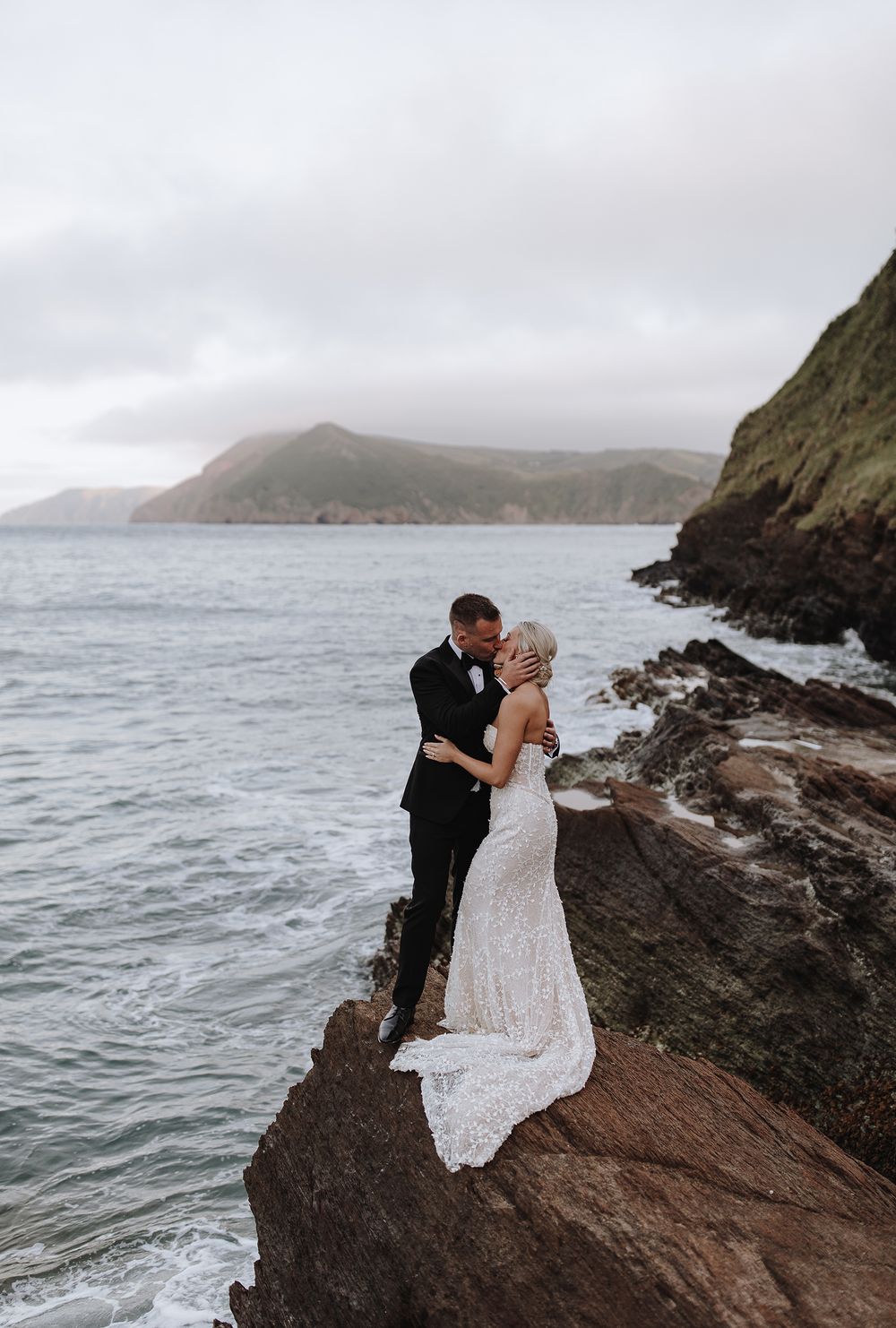 A romantic wedding couple embraces on dramatic coastal cliffs overlooking misty ocean waters.
