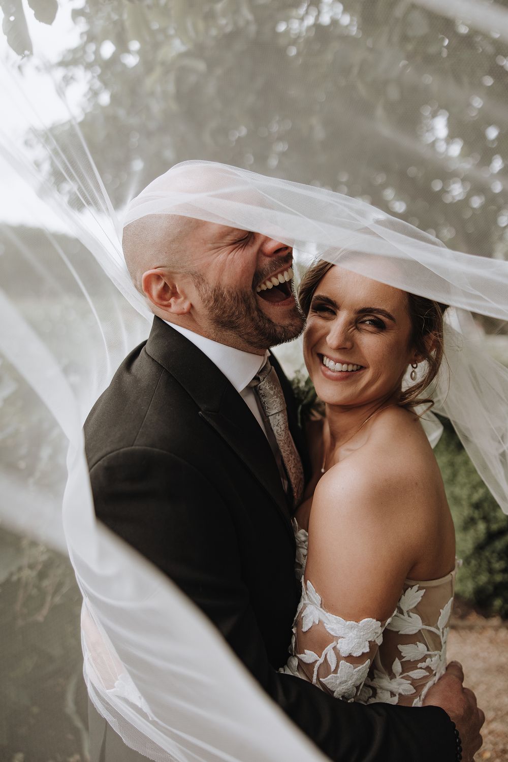 A joyful wedding couple shares an intimate moment under a flowing white veil.