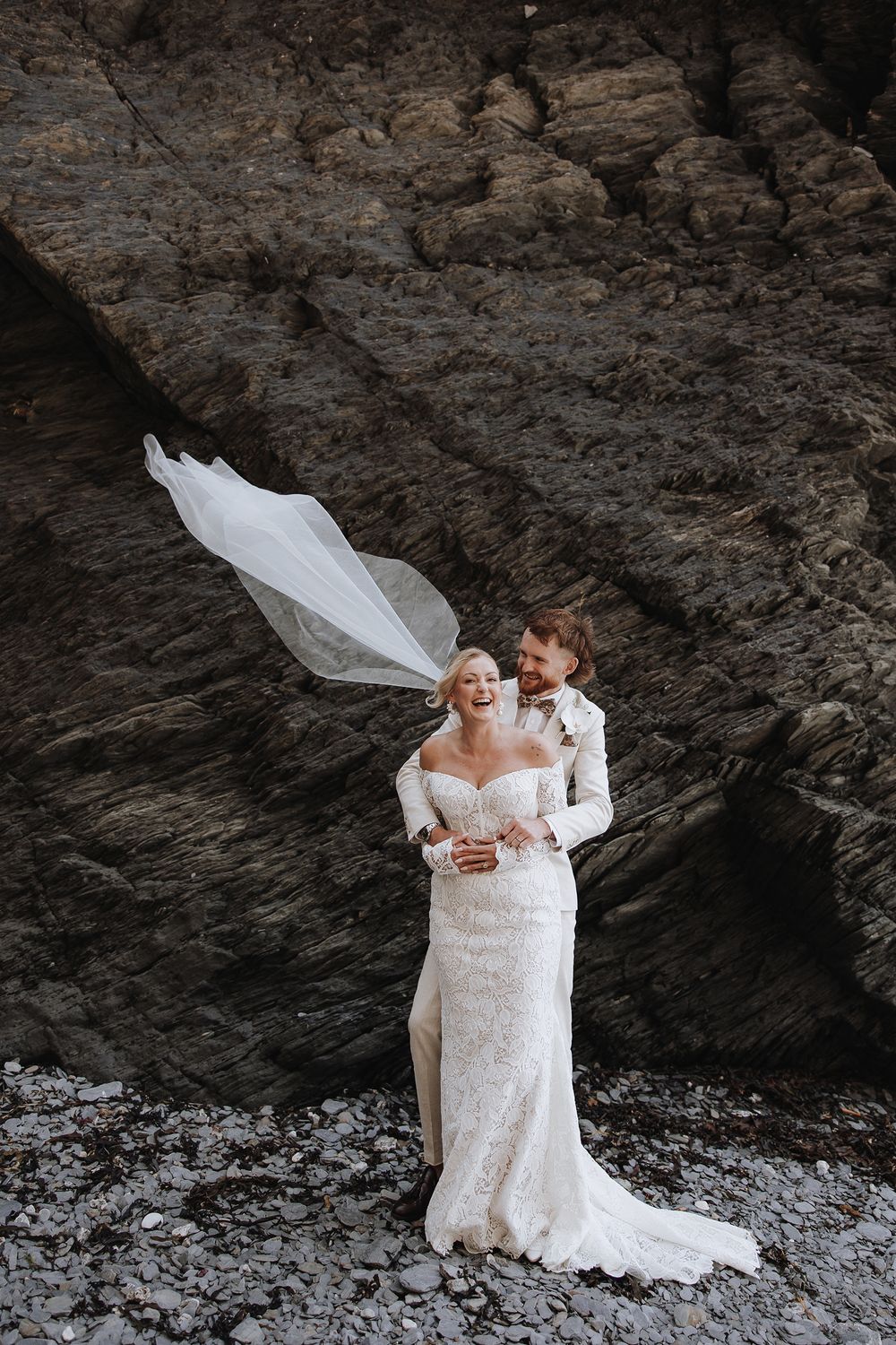 A couple in white wedding attire embraces under a rocky cliff overhang in a dramatic outdoor setting.