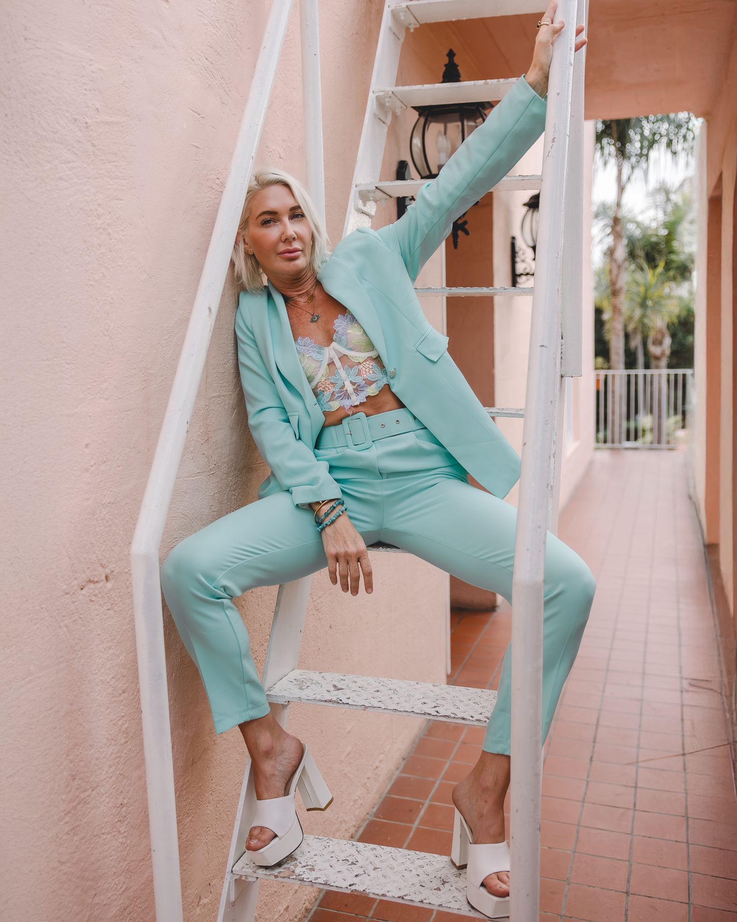 Model in mint green pantsuit and crop top poses against pink wall on tiled patio with white railing.