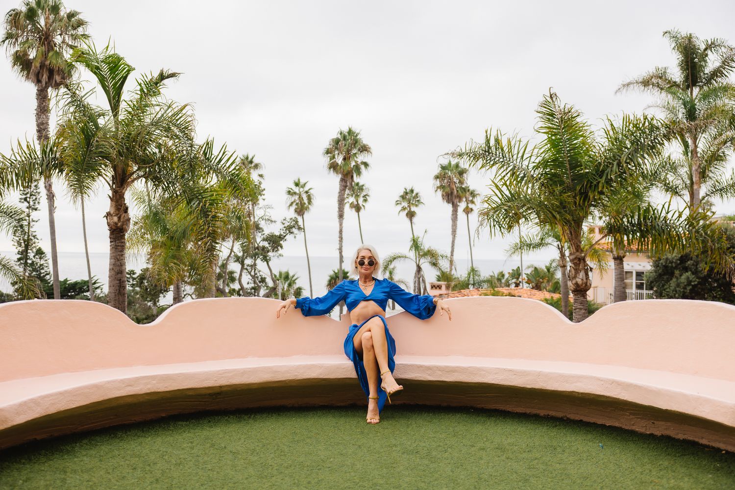 Someone in a blue dress sits on a curved pink wall with palm trees and green turf visible at a tropical location.