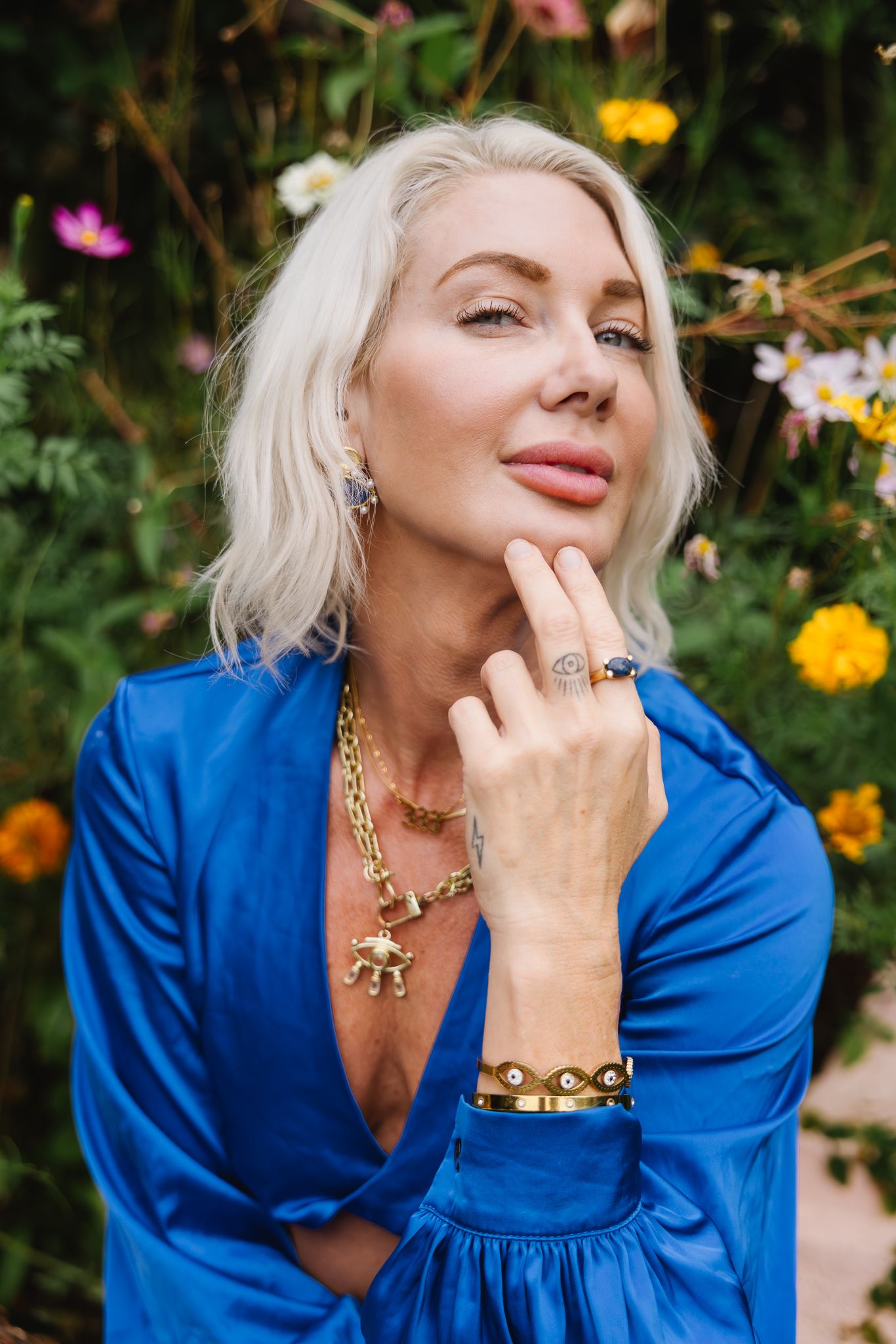 A person in a vibrant blue dress poses thoughtfully among colorful garden flowers.
