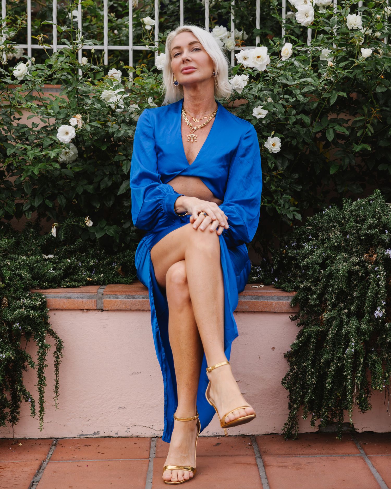 A person in a vibrant blue dress sits on a pink stone bench surrounded by lush white roses in a garden setting.