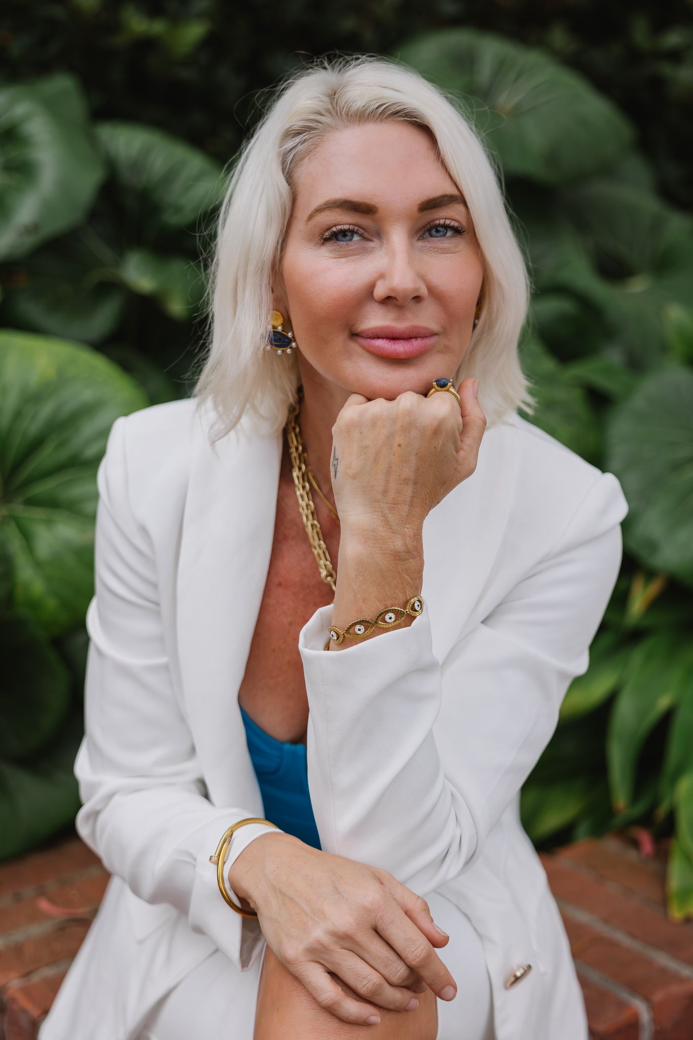 Elegant white blazer styled with gold chain accessories against lush green garden backdrop.