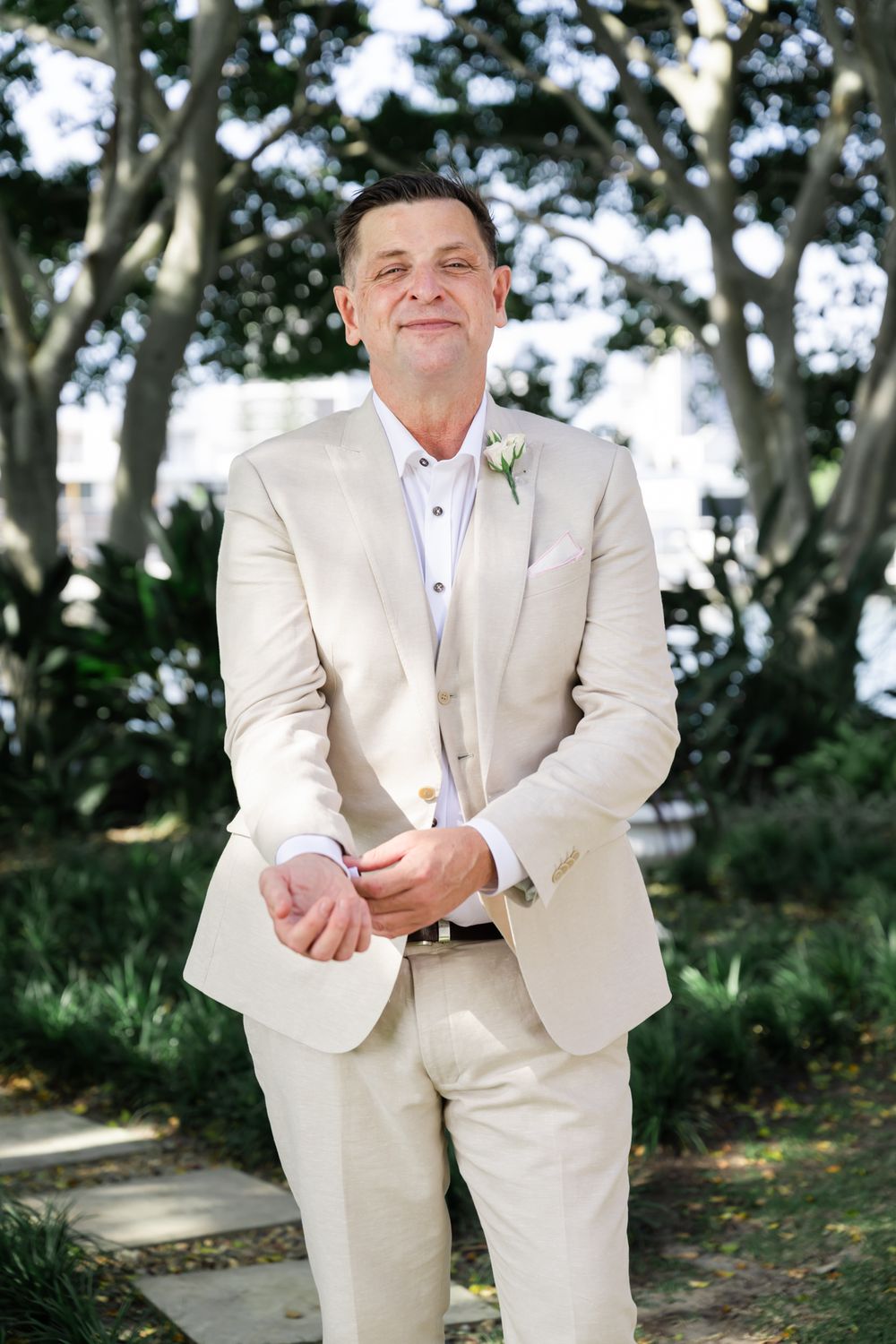 A person in a light beige suit sits on a bench in an outdoor garden setting with trees in the background.
