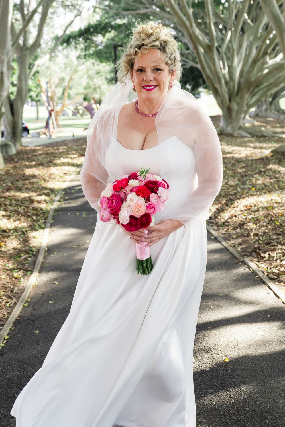 Bride in flowing white dress holds vibrant pink and red bouquet while standing on garden path in dappled sunlight.