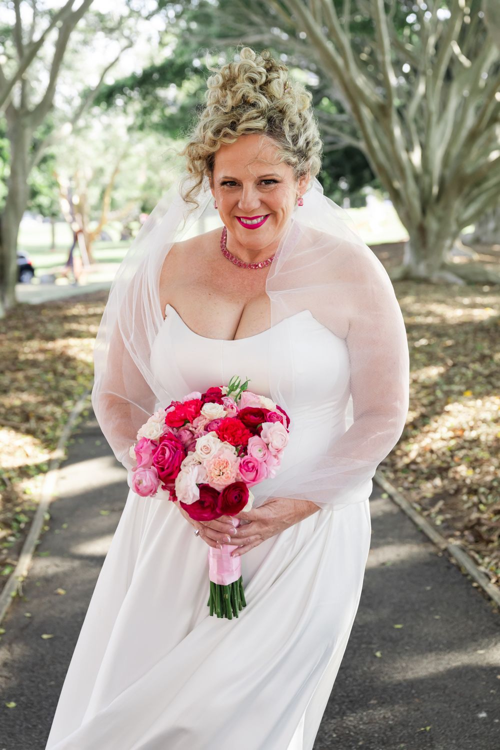 A bride in a white dress with sheer sleeves holds a vibrant pink and red bouquet while standing outdoors.