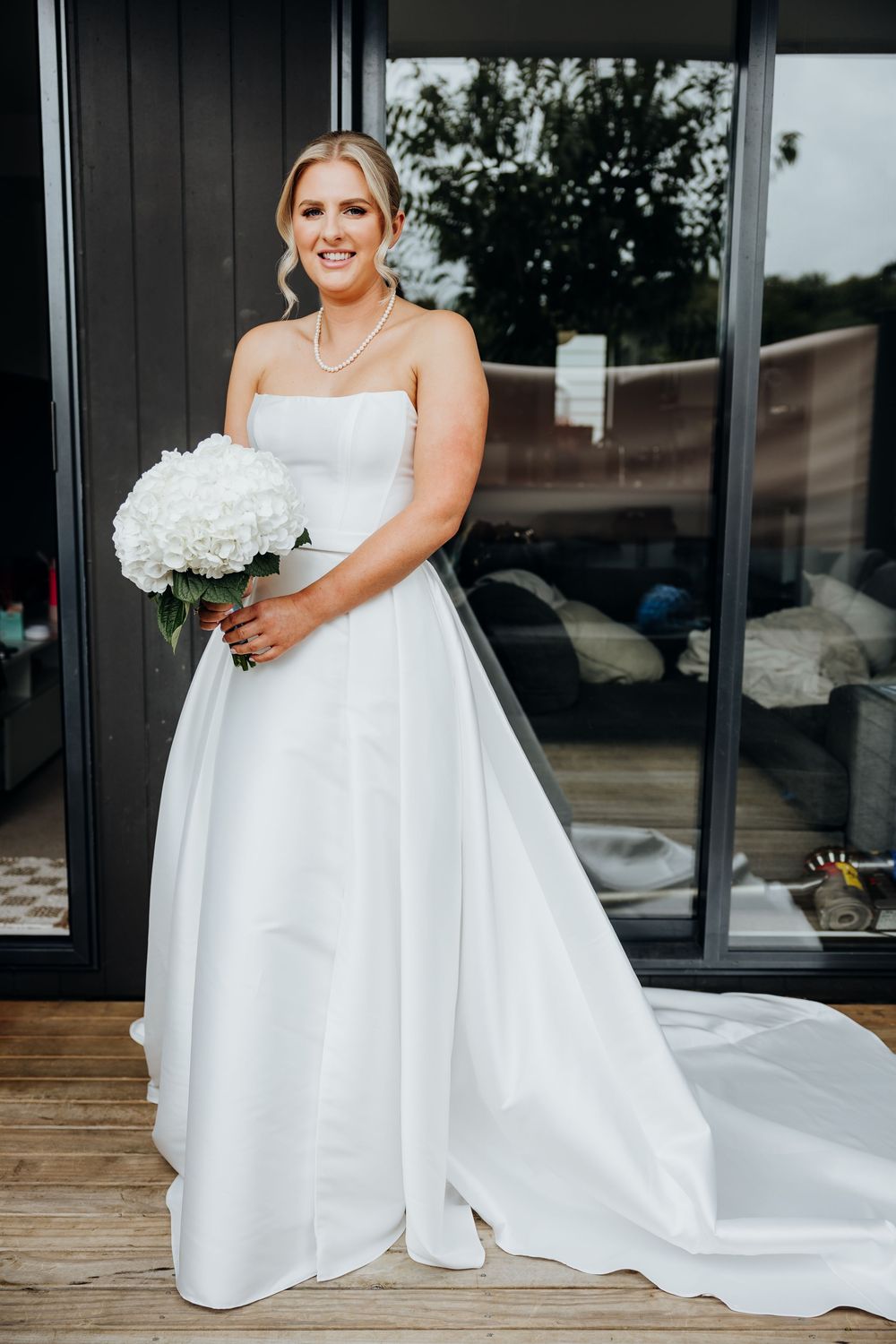 Bride in elegant white wedding gown poses on wooden deck with white floral bouquet.