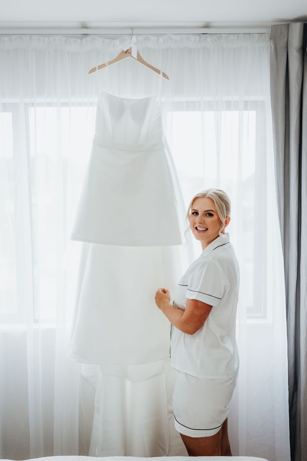 White wedding dress hanging by window with soft natural light streaming through sheer curtains.