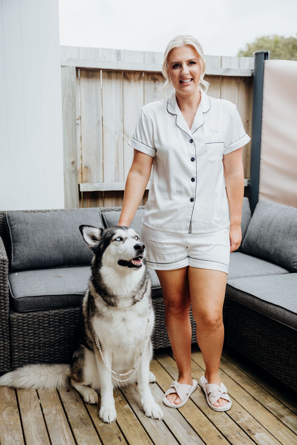 A Husky dog sits next to someone wearing white pajamas on an outdoor patio with grey furniture.