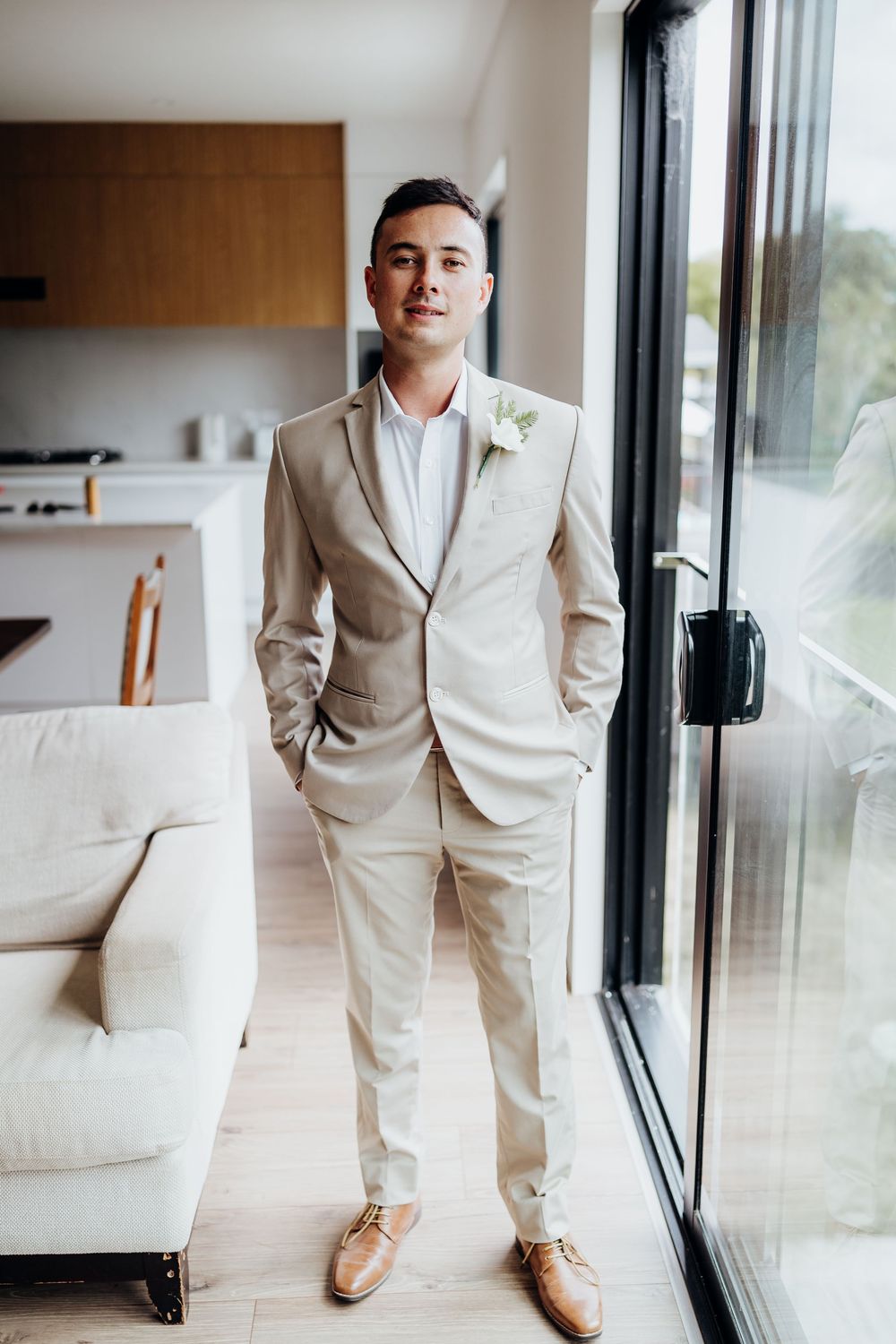 Groom in light beige suit and brown shoes poses by window in modern interior.