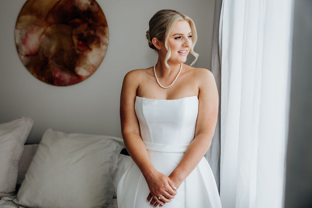 A bride in a white strapless dress sits by a window with elegant decor.
