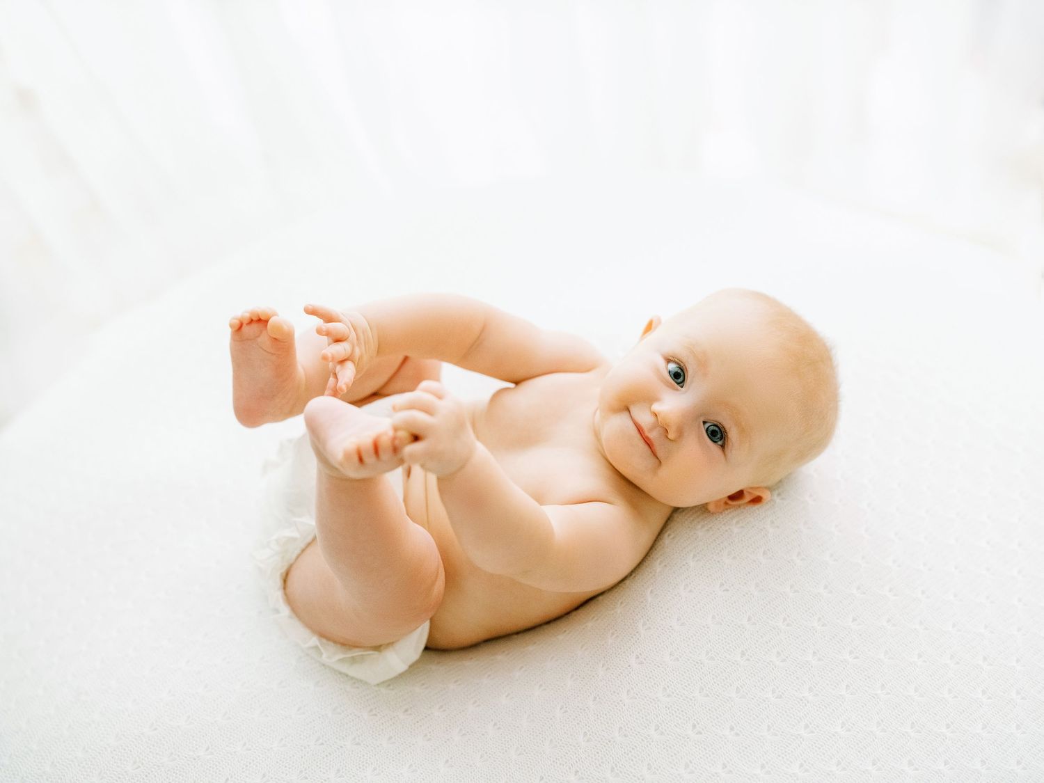 Adorable naked baby lying on white background playfully kicking legs in the air.