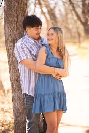A couple embraces near a tree trunk in a sunlit forest setting, with the woman wearing a blue dress.