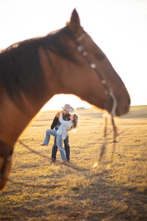 Person in light blue shirt bends down near brown horse in golden sunlit field at sunset.