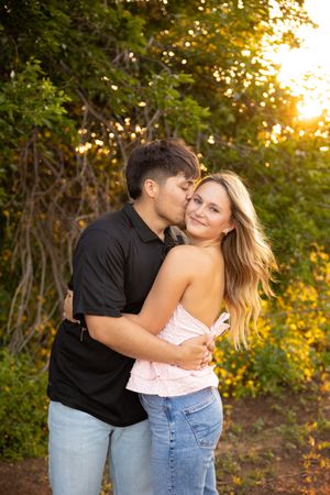 A couple embraces and kisses during a romantic sunset photoshoot in a natural outdoor setting.