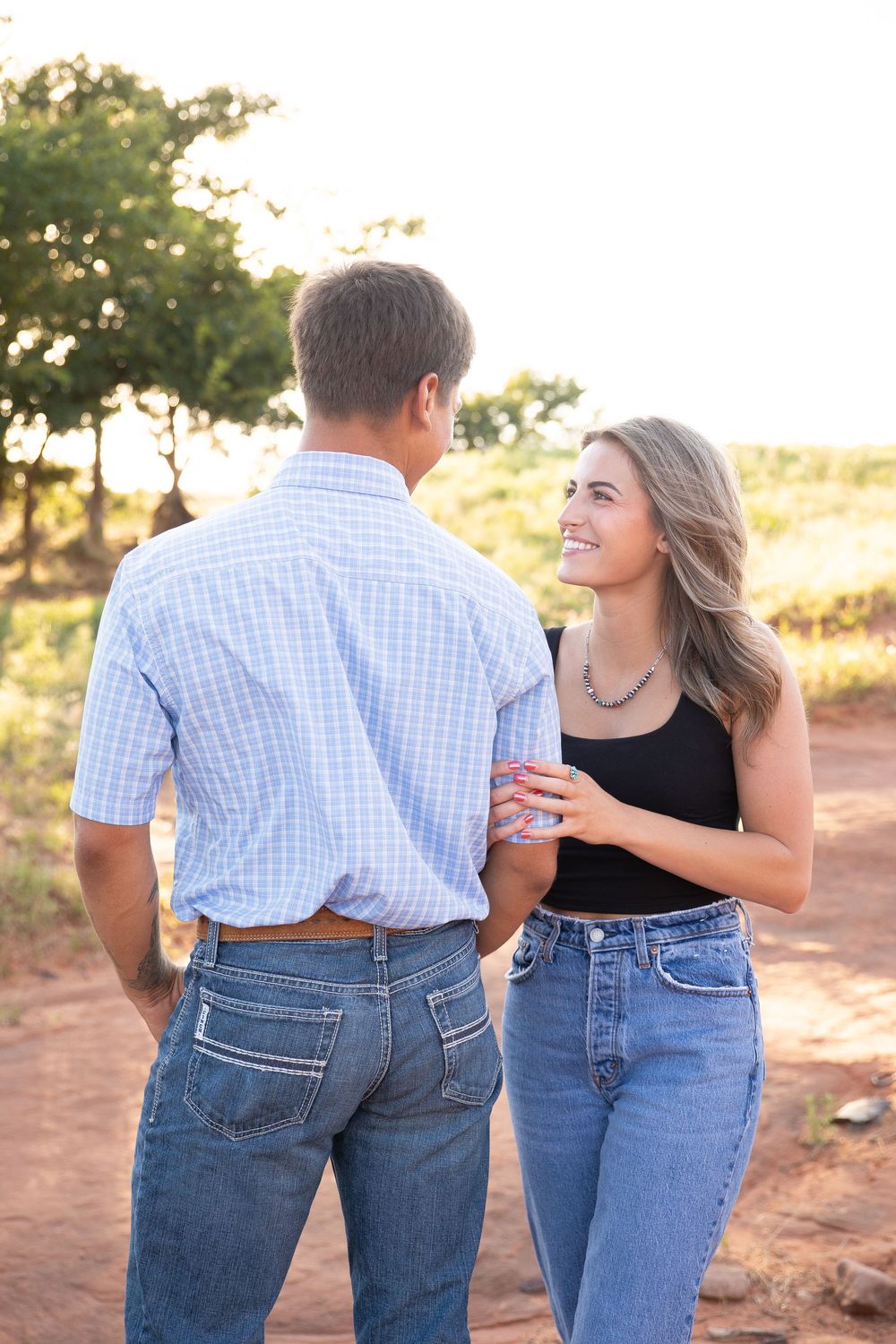 A couple stands together on a dirt path at sunset, wearing jeans and sharing a romantic moment in golden light.
