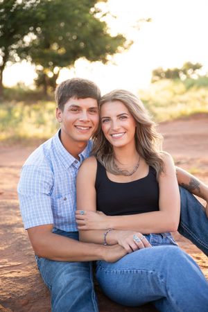 A couple sits close together on a dirt path during golden hour sunset with trees in the background.