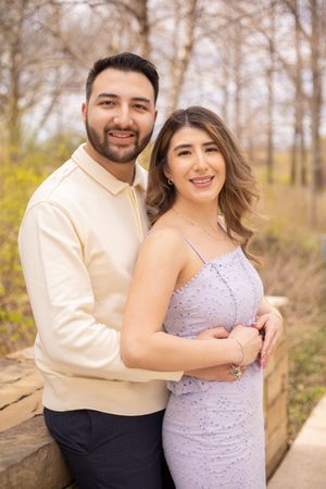 A couple poses outdoors in spring, wearing a cream colored shirt and lavender dress among bare trees.