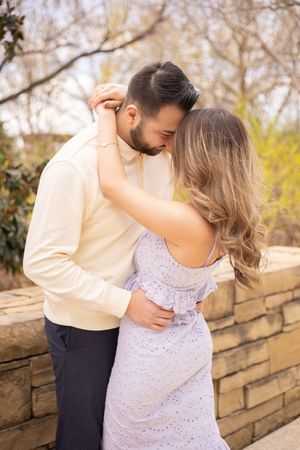 A romantic couple embraces against a stone wall backdrop with bare trees and yellow spring foliage in the background.