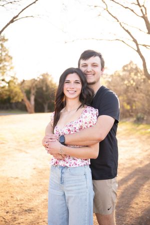 A couple embraces outdoors during golden hour with bare trees and sunlit field in the background.