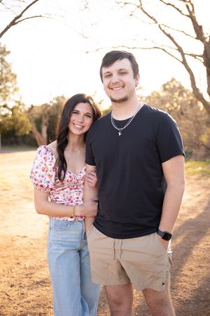 A couple poses together outdoors at sunset wearing casual clothing and smiling warmly at the camera.