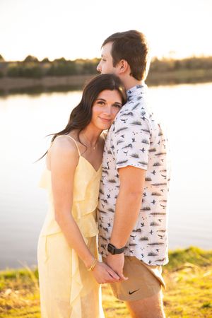A couple stands together at sunset by a lake, with the person in yellow embracing their partner from behind.