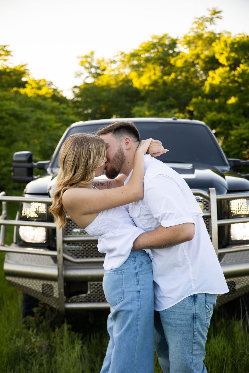 A romantic couple embraces by a pickup truck at sunset in a grassy field.