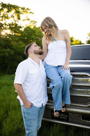 Romantic couple poses against silver truck grille during sunset photoshoot in grassy field.